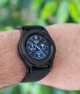 A close-up of an arm wearing a smart watch with a black strap, displaying various health and activity metrics including a step counter and heart rate monitor, set against a blurred green leaf background.