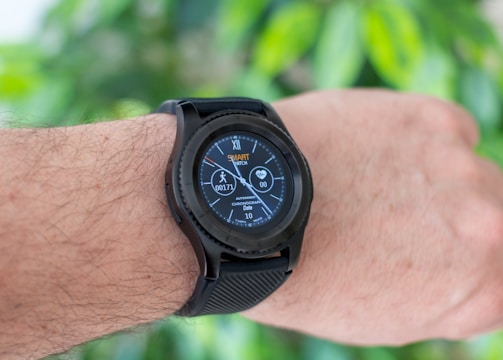 A close-up of an arm wearing a smart watch with a black strap, displaying various health and activity metrics including a step counter and heart rate monitor, set against a blurred green leaf background.