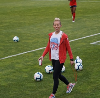 A person in athletic clothing walks across a grass field, holding a drink can and a skipping rope. Several soccer balls are scattered on the field. Other individuals in red athletic attire are in the background, suggesting a sports practice or event.