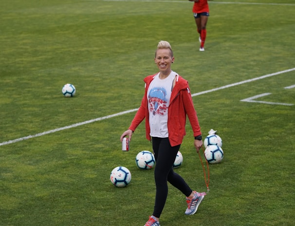 A person in athletic clothing walks across a grass field, holding a drink can and a skipping rope. Several soccer balls are scattered on the field. Other individuals in red athletic attire are in the background, suggesting a sports practice or event.