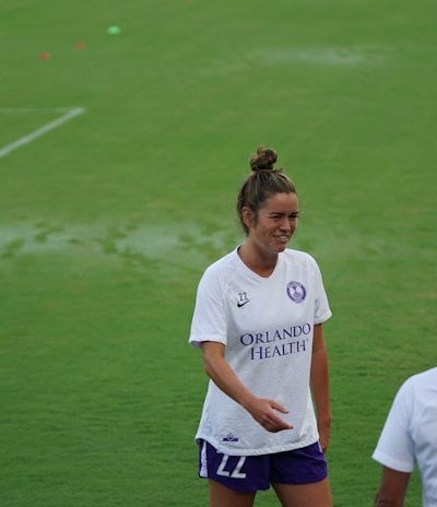 A woman wearing a white sports jersey with 'Orlando Health' written on it stands on a grassy field, smiling and engaging with another person who is partially visible. She has her hair tied up and is wearing shorts. The field is well-maintained and appears to be an athletic or sports setting.