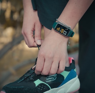 A person is tying the laces of a black, turquoise, and pink sneaker. They are wearing a digital watch that displays health tracking information on their wrist, along with a bracelet. The background is blurred, suggesting an outdoor setting.
