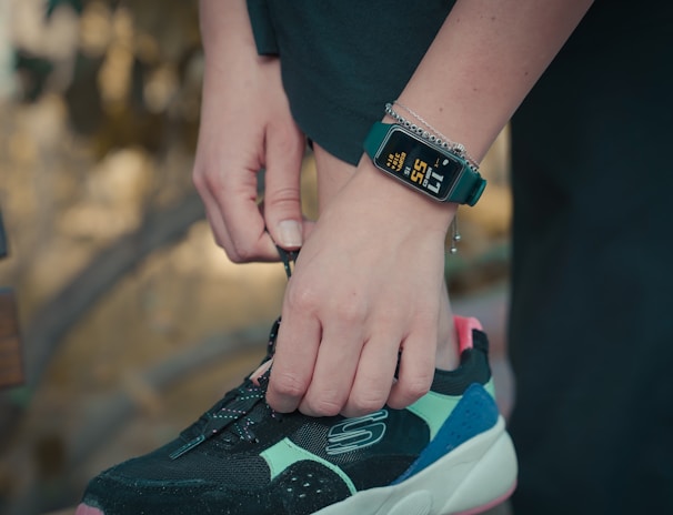 A person is tying the laces of a black, turquoise, and pink sneaker. They are wearing a digital watch that displays health tracking information on their wrist, along with a bracelet. The background is blurred, suggesting an outdoor setting.