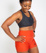 A smiling woman wearing a dark sports bra and bright orange athletic shorts with a logo on the side. She stands with her hands on her hips, exuding confidence and athleticism. Her hair is pulled back, and she seems to be in a positive mood.