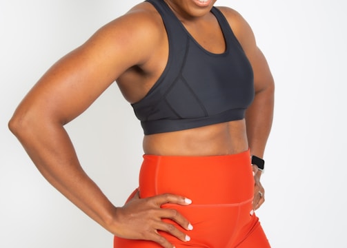 A smiling woman wearing a dark sports bra and bright orange athletic shorts with a logo on the side. She stands with her hands on her hips, exuding confidence and athleticism. Her hair is pulled back, and she seems to be in a positive mood.