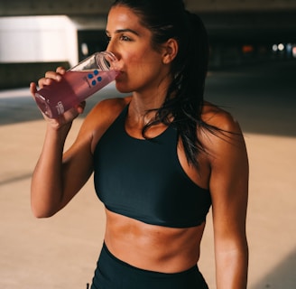 A fit woman is standing in a dimly lit space, wearing a black sports bra and shorts. She is holding a pink drink in a transparent bottle and is in the act of taking a sip. Her hair is pulled back, and she wears a watch on her left wrist.