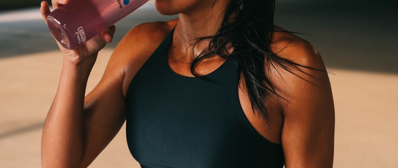 A fit woman is standing in a dimly lit space, wearing a black sports bra and shorts. She is holding a pink drink in a transparent bottle and is in the act of taking a sip. Her hair is pulled back, and she wears a watch on her left wrist.