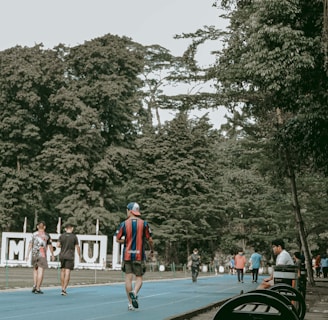 A vibrant outdoor scene featuring a running track surrounded by lush green trees. People are actively walking or jogging on the blue track, some dressed in sportswear. Benches and a decorative sculpture are visible alongside the track. The setting appears lively and recreational.