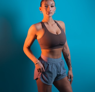 A person stands confidently against a solid turquoise background, wearing a brown sports bra and gray-blue athletic shorts. The individual has a fit physique and is adorned with earrings and a bracelet. Tattoos are visible on their arm, adding to their sporty appearance.