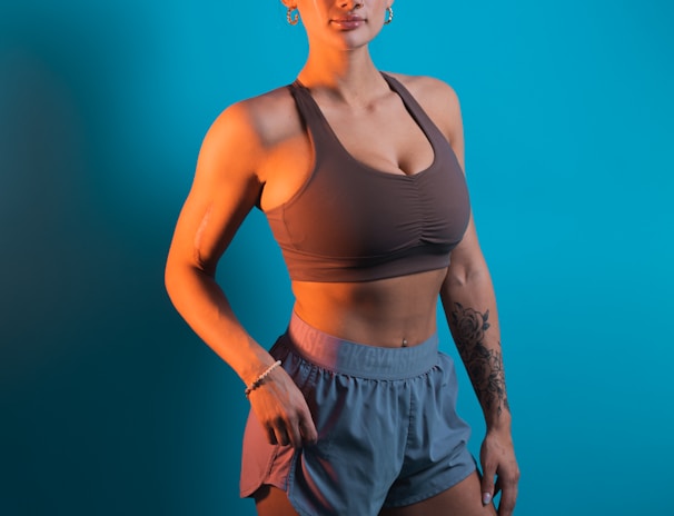 A person stands confidently against a solid turquoise background, wearing a brown sports bra and gray-blue athletic shorts. The individual has a fit physique and is adorned with earrings and a bracelet. Tattoos are visible on their arm, adding to their sporty appearance.