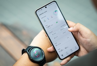 A person holds a smartphone displaying the Samsung Health app, showing metrics such as daily activity, steps, calories, and exercise data. The individual also wears a smartwatch on their wrist, which has a colorful display indicating health statistics.