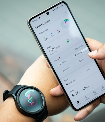 A person holds a smartphone displaying the Samsung Health app, showing metrics such as daily activity, steps, calories, and exercise data. The individual also wears a smartwatch on their wrist, which has a colorful display indicating health statistics.