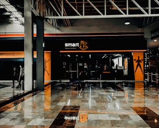 A fitness center entrance with the branding 'smart fit' illuminated above the doors. The interior has a modern design, featuring dark glass and bright orange accents. A reflective marble floor extends in front, capturing reflections from the surroundings. There are two individuals standing and walking towards the left.