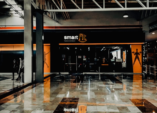 A fitness center entrance with the branding 'smart fit' illuminated above the doors. The interior has a modern design, featuring dark glass and bright orange accents. A reflective marble floor extends in front, capturing reflections from the surroundings. There are two individuals standing and walking towards the left.